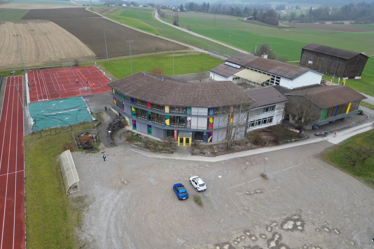 Zum Schulhaus mit Turnhalle und Aula kommt ein Neubau. Dieser wird am (im Bild unteren) Rand des Kiesplatzes bei der 100-Meter-Anlage platziert.