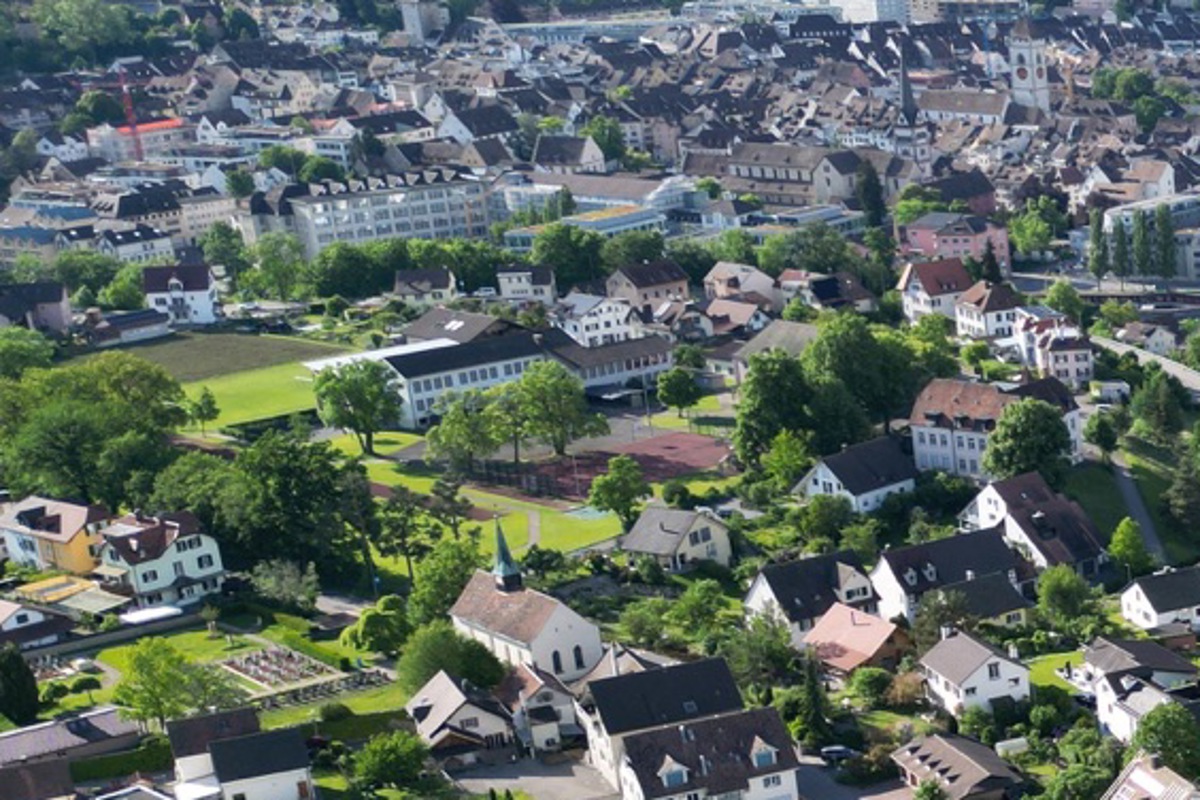 Auf dem Schulareal Spilbrett (Bildmitte, mit viel Wiese rundum) plant die Gemeinde Feuerthalen eine neue Doppelturnhalle. Vorgesehen ist, dass der heutige rote Platz dem Neubau weicht, während die links im Bild als dunkler Acker sichtbare Parzelle 2000 zur Mitfinanzierung verkauft werden soll.